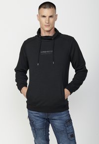 Sudadera negra con capucha y textura de espiga, con cordón, logo en el pecho y puños acanalados; combinada con pantalones cargo azules.