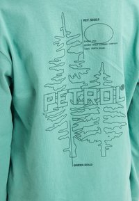 Mintgrünes Langarmshirt mit einem Baumgrafik und dem Text "PETROL" in Dunkelgrün. Blattmuster zieren das Design.