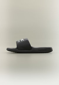 Black slide sandal with a wide strap, made of smooth synthetic material. Features white Lacoste logo and textured sole for grip.
