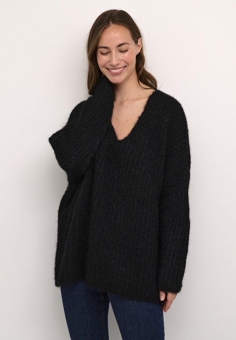 Maglione nero a coste con scollatura a V profonda, vestibilità oversized e maniche lunghe. Tessuto a maglia con un aspetto morbido e peloso. Abbinato a jeans blu scuro.
