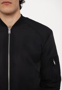 Homme portant une veste bomber noire avec une fermeture éclair argentée sur le devant et une poche zippée sur la manche supérieure, sur fond blanc.