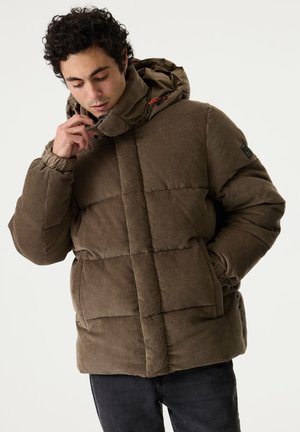 Uomo con capelli ricci indossa un piumino marrone in velluto a coste con cappuccio, guarda verso il basso mentre tiene il colletto, su uno sfondo semplice.