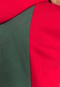 Felpa con cappuccio caratterizzata da un design a blocchi di colore, con tessuto rosso e verde scuro, texture liscia e cuciture curve per un aspetto moderno.