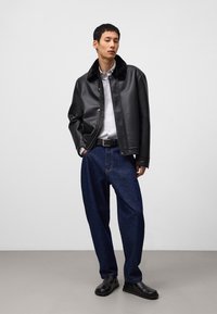 Svart fuskpälsjacka med krage av päls, vit skjorta med knappar, blå baggy jeans och svarta slip-on skor, poserad mot en neutral bakgrund.