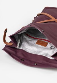 Borsa impermeabile color mogano con dettagli in pelle color tan e chiusure a scatto, dotata di un interno a quadri grigi e una tasca con zip rossa.