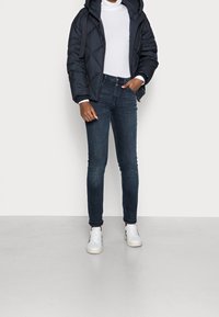 Person som bär en svart vadderad jacka, vit turtleneck, mörkblå jeans och vita sneakers, stående med en hand i fickan.