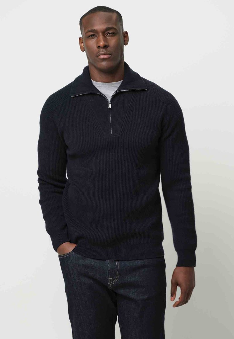 Granatowy sweter prążkowany z ćwierć zamkiem, z V-kształtnym wykończeniem przy szyi. Połączony z ciemnymi dżinsami. Luźny krój.