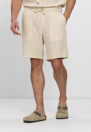 Mand iført beige snørede shorts og beige slip-on træsko, stående med den ene hånd i lommen og benene synlige under knæet.