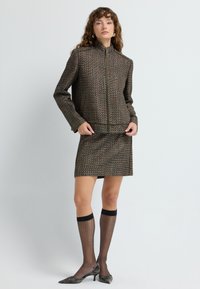 Veste marron texturée avec un col haut et une fermeture éclair, assortie d'une jupe coordonnée. Des chaussettes noires montantes et des chaussures à bout pointu complètent le look.