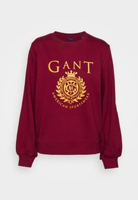 Sudadera color burdeos con puños y cuello elásticos; presenta un gran logo amarillo con un diseño de corona y laurel, que dice "GANT AMERICAN SPORTSWEAR."