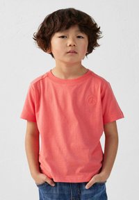 T-shirt corail à manches courtes en coton doux, avec un col côtelé et un logo subtil sur la poitrine, associé à un jean en denim.