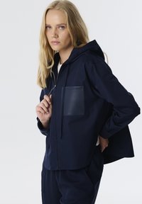 Chaqueta con capucha de color azul marino, hecha de tela suave, con un bolsillo frontal, cierre de cremallera y corte holgado. Detalles texturizados en el bolsillo.