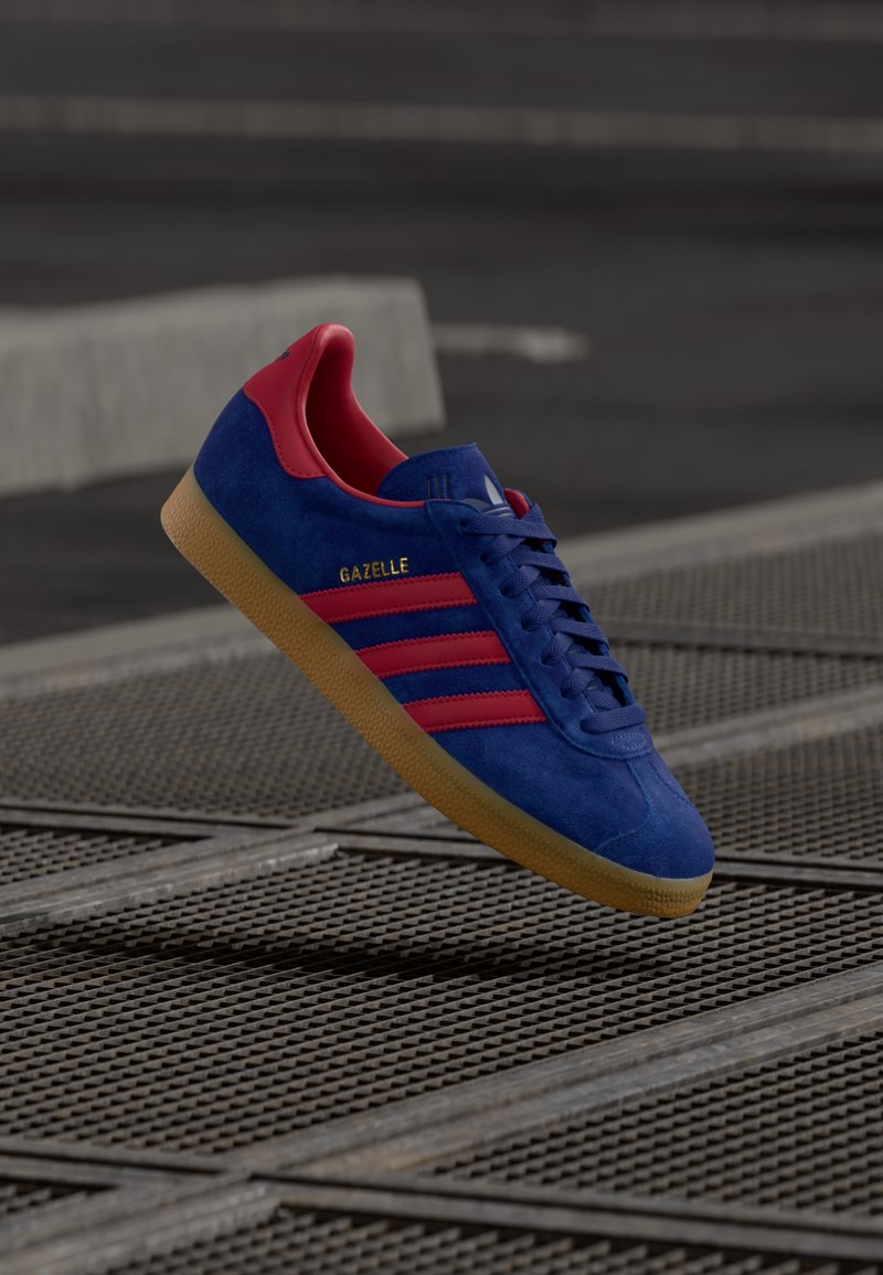 Tênis Adidas Gazelle azul com listras vermelhas e calcanhar, sola de borracha, flutuando acima de uma superfície de grade metálica em ambiente urbano.