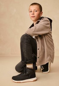 Czarne sneakersy typu high-top z gumową podeszwą, teksturowanymi bocznymi panelami i wiązanym zapięciem. Uzupełnione szarymi dżinsami i beżową kurtką z prążkowanej tkaniny.