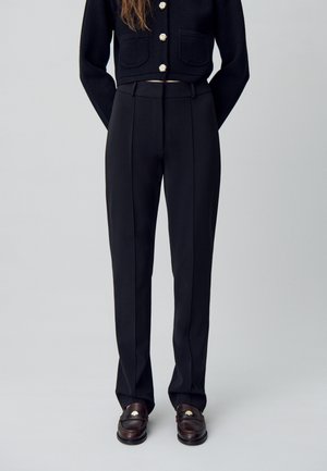 Pantalon noir cintré avec une taille haute et des plis marqués, associé à des mocassins sombres avec un accent doré.