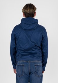 Giacca con cappuccio blu navy realizzata in tessuto leggero, progettata con una chiusura frontale con cerniera e polsini elasticizzati, abbinata a jeans in denim.