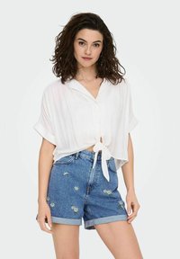 Blouse blanche à manches courtes et à boutons, avec un devant noué ; associée à un short en denim bleu avec broderies florales.