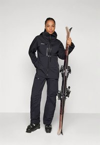 Strój narciarski składający się z czarnej wodoodpornej kurtki i spodni z zamkami błyskawicznymi, zestawiony z czarnymi butami narciarskimi i nartami, stojący na jednolitym tle.