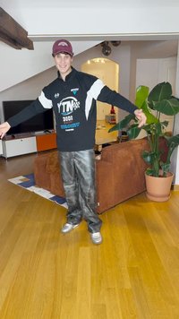 Juodas sportinis džemperis su baltais ir mėlynais akcentais, pilkos džinsinio audinio kelnes ir sidabriniai sportbačiai, esant ryškiai patalpoje su augalais.