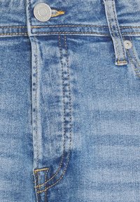 Vaaleansininen farkkukangas, jossa on pehmeä rakenne, kontrastiväriset ompeleet ja hopea nappi vyötäröllä.