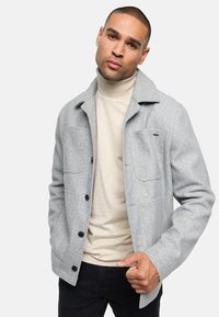 Veste en laine grise avec un design à col, des poches avant et des boutons noirs, portée sur un col roulé beige et un pantalon noir.