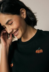 T-shirt noir avec broderie de citrouille en sequins orange sur la poitrine. L'encolure est côtelée et le tissu semble doux et confortable.