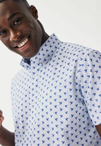 Camicia a maniche corte in tessuto bianco con stampa di uccelli blu. Presenta un colletto con bottoni e una tasca sul petto. Texture liscia.