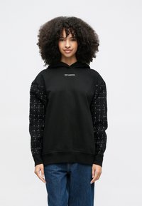 Γυναίκα με σγουρά μαλλιά, φορώντας μαύρο φούτερ με μοτίβα στα μανίκια και το κείμενο «KARL LAGERFELD», σε συνδυασμό με μπλε τζιν, στέκεται μπροστά σε λευκό φόντο.