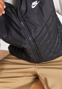 Svart pufferväst med dragkedja fram, med en kviltad textur och en vit Nike-logotyp på övre vänstra sidan. Åtföljd av beiga byxor.