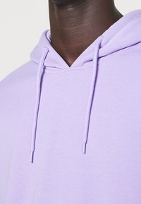 Světle fialová mikina s kapucí, která má dvojitou podšívku, šňůrku s kovovými konci a měkkou, texturovanou tkaninu. Viditelné švy kolem krku.