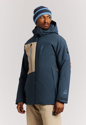 Man die een blauwe en beige winterjas met capuchon en een blauw gestreepte gebreide muts draagt en opzij kijkt tegen een effen achtergrond.