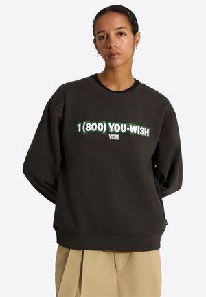 Giovane con capelli corti indossa una felpa nera con la scritta "1 (800) YOU-WISH VANS" e pantaloni beige su sfondo neutro.