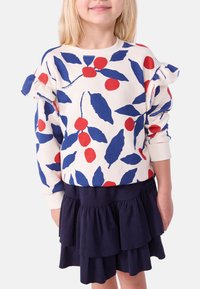 Weißer Sweatshirt mit blauem und rotem Blatt- und Kirschmuster, mit Rüschenschultern. Kombiniert mit einem dunkelblauen, gestuften Rock.