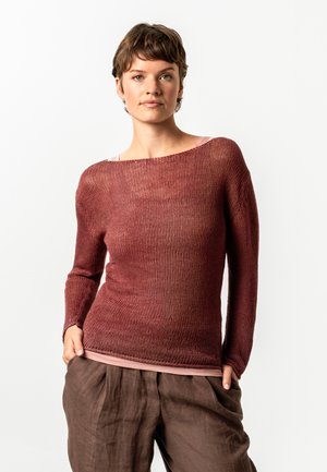 Woman with short brown hair wears a loose, long-sleeve, rust-colored knit sweater over a pink top and brown wide-leg pants, posed with hands in pockets.