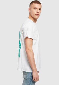 Hvid bomulds t-shirt med fede tealfarvede og blå grafiske bogstaver på ryggen, korte ærmer og en afslappet pasform. Båret med lyseblå jeans.