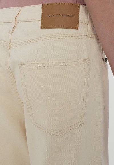Pantaloni din denim bej cu o etichetă din piele marcata "TIGER OF SWEDEN," având un design standard al buzunarului din spate și detalii de cusătură subtile.