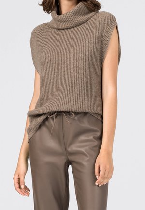 Kvinde iført ærmeløs, ribstrikket, rullekrave-sweater i taupe med brune læderbukser med snørre.