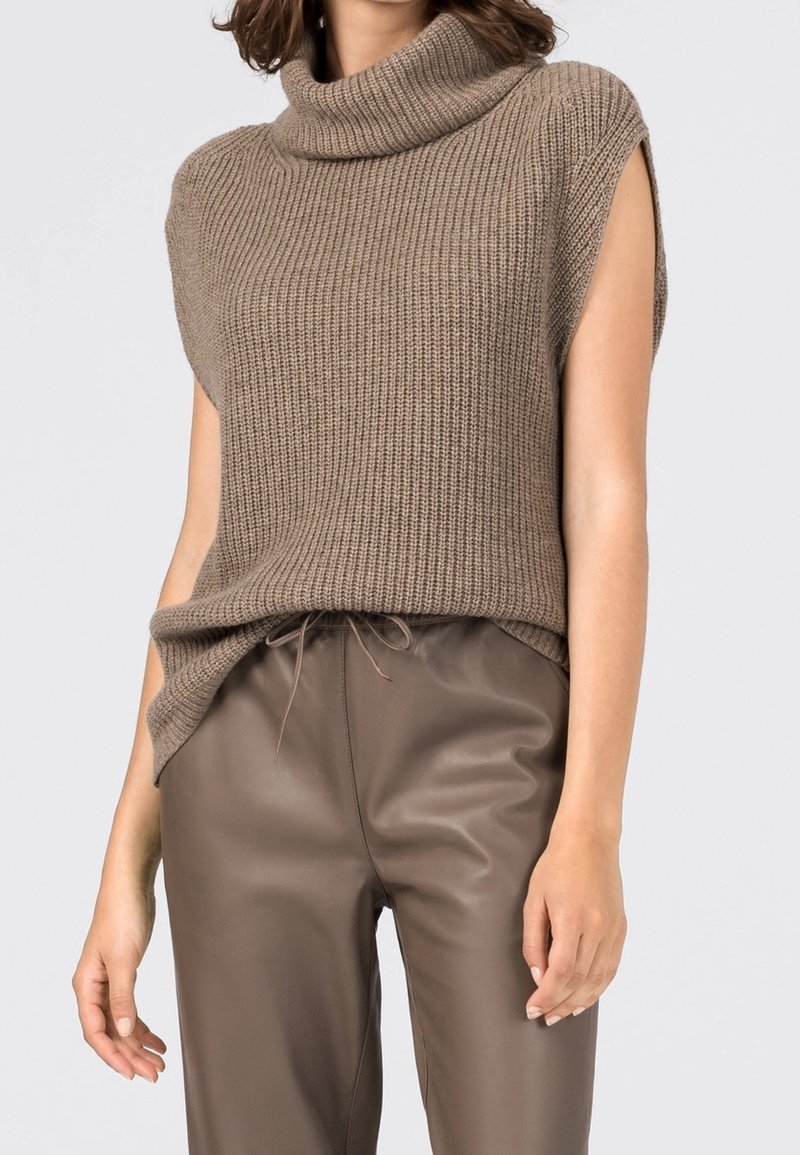 Femme portant un pull sans manches côtelé à col roulé taupe avec un pantalon en cuir marron à cordon.