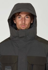 Grå isolerad jacka med hög krage och stora bröstfickor. Har en huva och textil med strukturerad yta samt kontrasterande mörka accenter.