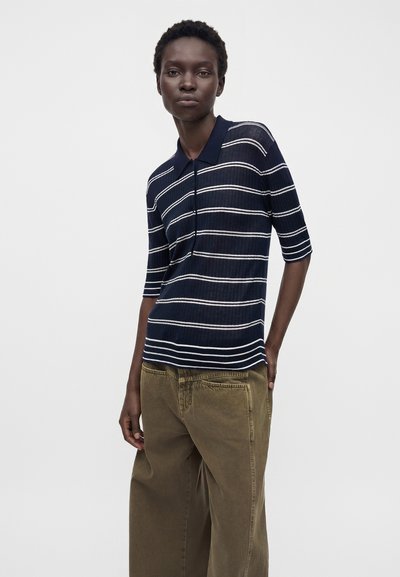 Femme portant un polo bleu marine à manches courtes avec des rayures blanches et un pantalon marron ample, debout sur un fond clair uni.