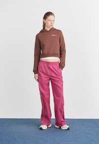 Sweatshirt castanho curto com logótipo branco, combinado com calças largas cor-de-rosa com riscas brancas nas laterais. Ténis neutros completam o visual.