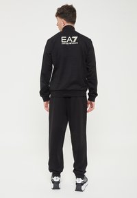 Giacca da tuta nera con collo alto, polsini elasticizzati e pantaloni abbinati. Logo "EA7 Emporio Armani" stampato sulla schiena. Tessuto liscio.