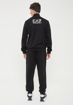 Giacca da tuta nera con collo alto, polsini elasticizzati e pantaloni abbinati. Logo "EA7 Emporio Armani" stampato sulla schiena. Tessuto liscio.