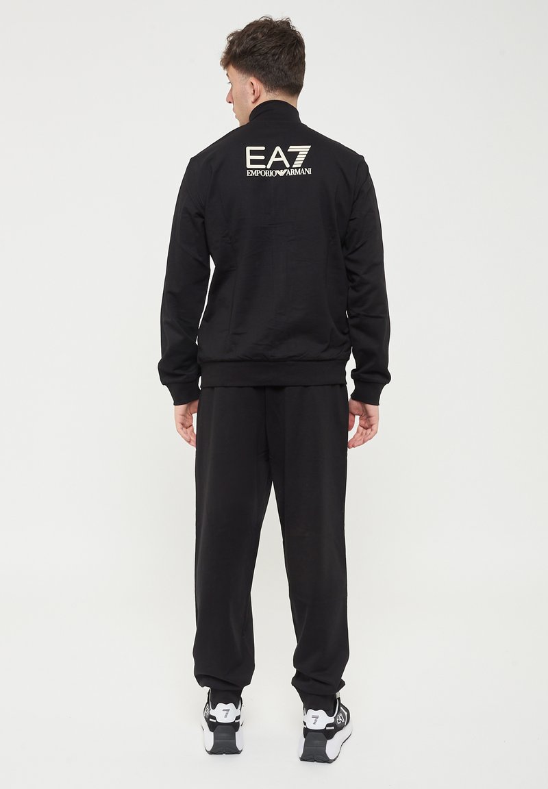 Giacca da tuta nera con collo alto, polsini elasticizzati e pantaloni abbinati. Logo "EA7 Emporio Armani" stampato sulla schiena. Tessuto liscio.