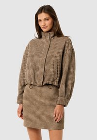 Brun houndstooth-jakke med høj krave og elastisk talje, parret med en matchende tætsiddende nederdel. Begge dele har et tekstureret stof.
