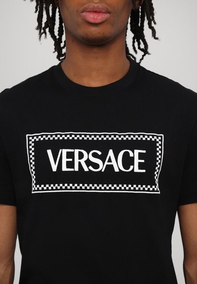 Melnas kokvilnas T-krekls ar baltu izšūtu "VERSACE" logotipu, ko ieskauj ruļļa raksts. Īsas piedurknes un apaļa kakla izgriezums.