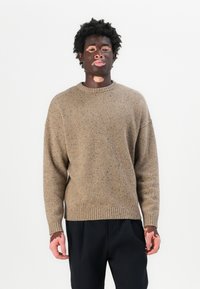 Maglione a maglia marrone macchiato con scollatura rotonda e polsini a costine. Il tessuto ha un aspetto tessurizzato. Indossato sopra pantaloni scuri.