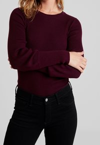 Burgunderfarbendes, geripptes Langarmoberteil mit Rundhalsausschnitt, kombiniert mit schwarzen, hoch taillierten Jeans mit Knopfverschluss und vorderen Taschen.