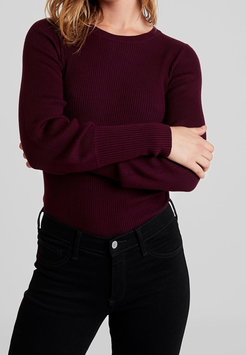Burgunderfarbendes, geripptes Langarmoberteil mit Rundhalsausschnitt, kombiniert mit schwarzen, hoch taillierten Jeans mit Knopfverschluss und vorderen Taschen.