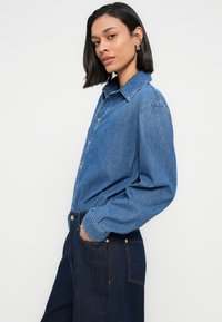 Μπλε πουκάμισο από τζιν με κουμπιά, γιακά, μακριά μανίκια και άνετη εφαρμογή. Φορεμένο με σκούρο μπλε τζιν παντελόνι, που δείχνει λεπτομέρεια τσέπης.